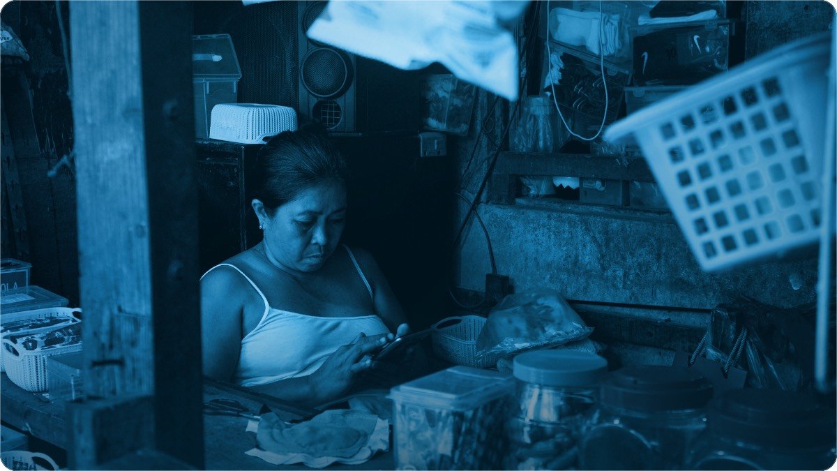A woman sitting alone inside her small shop, looking down at something she is holding.