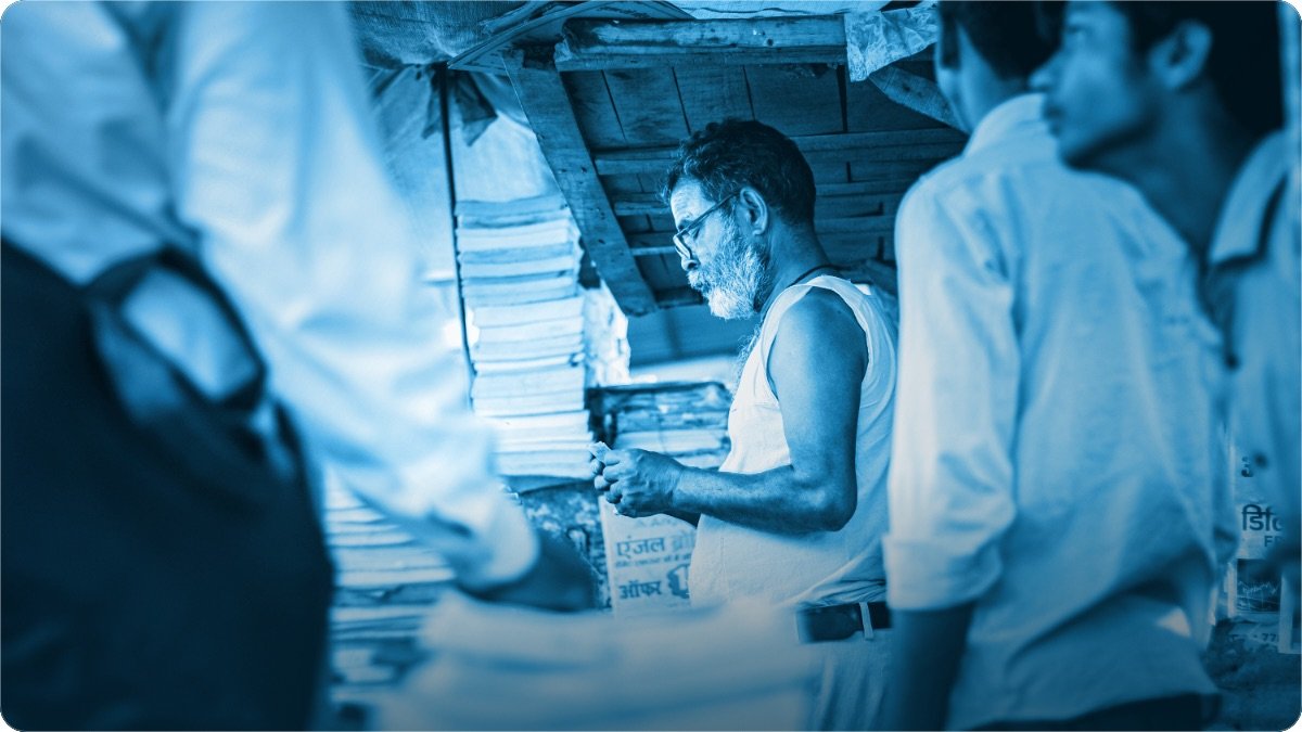 Dukandar apne chhote se khoke par paise gin raha hai aur customer counter ke gird khare hain.