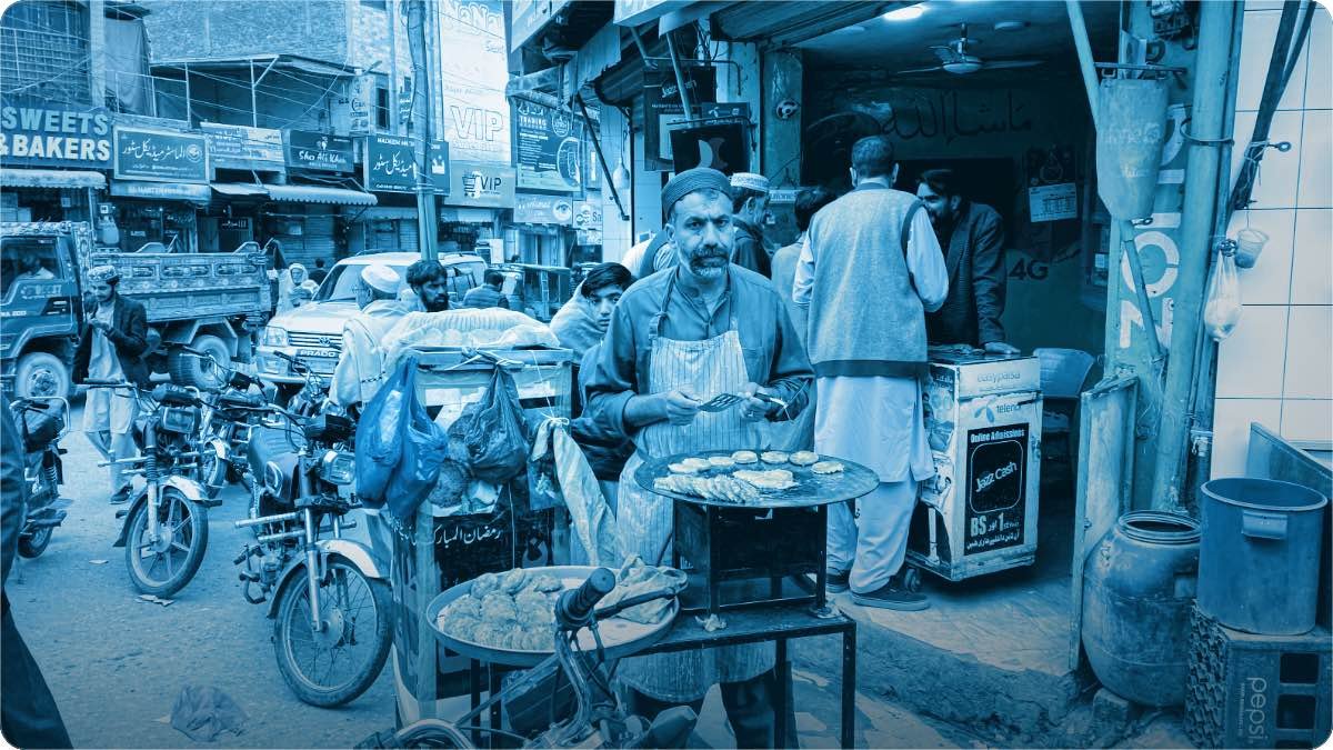 Bhara hua bazaar, thele par khana banata hua banda, khari motorcyclein aur dono taraf khuli dukanein.