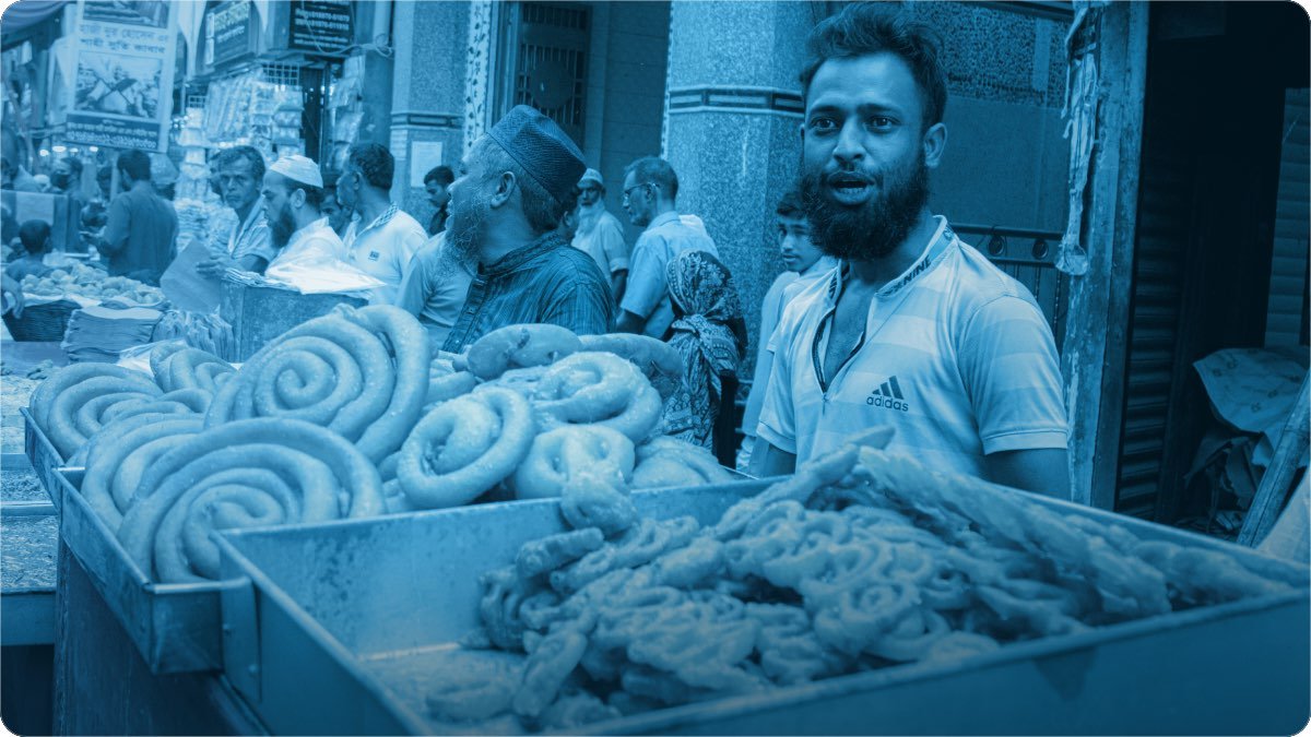 A busy street sweet stall, trays of fresh sweets in front and customers crowding the shops behind.
